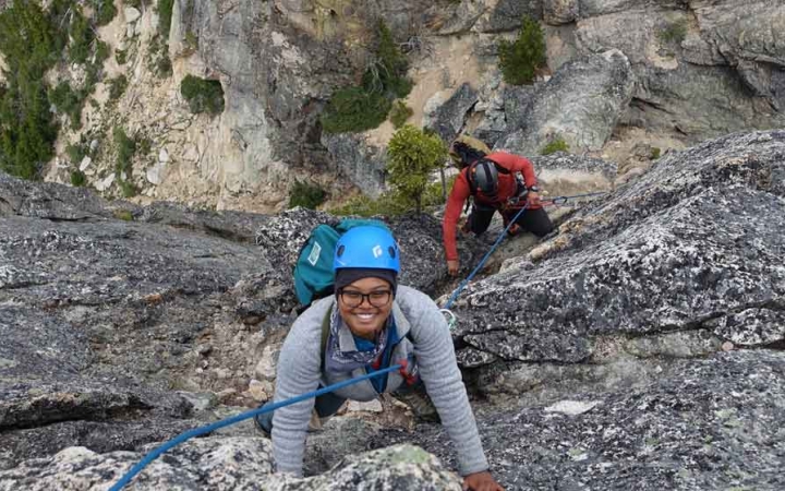 a person looks up and smiles at the camera while rock climbing. another person climbs below her. 